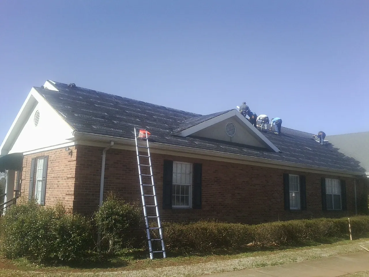 Crew tearing shingles off a brick ranch roof with a ladder set — Emergency Roof Repair work by our crew in Mayodan