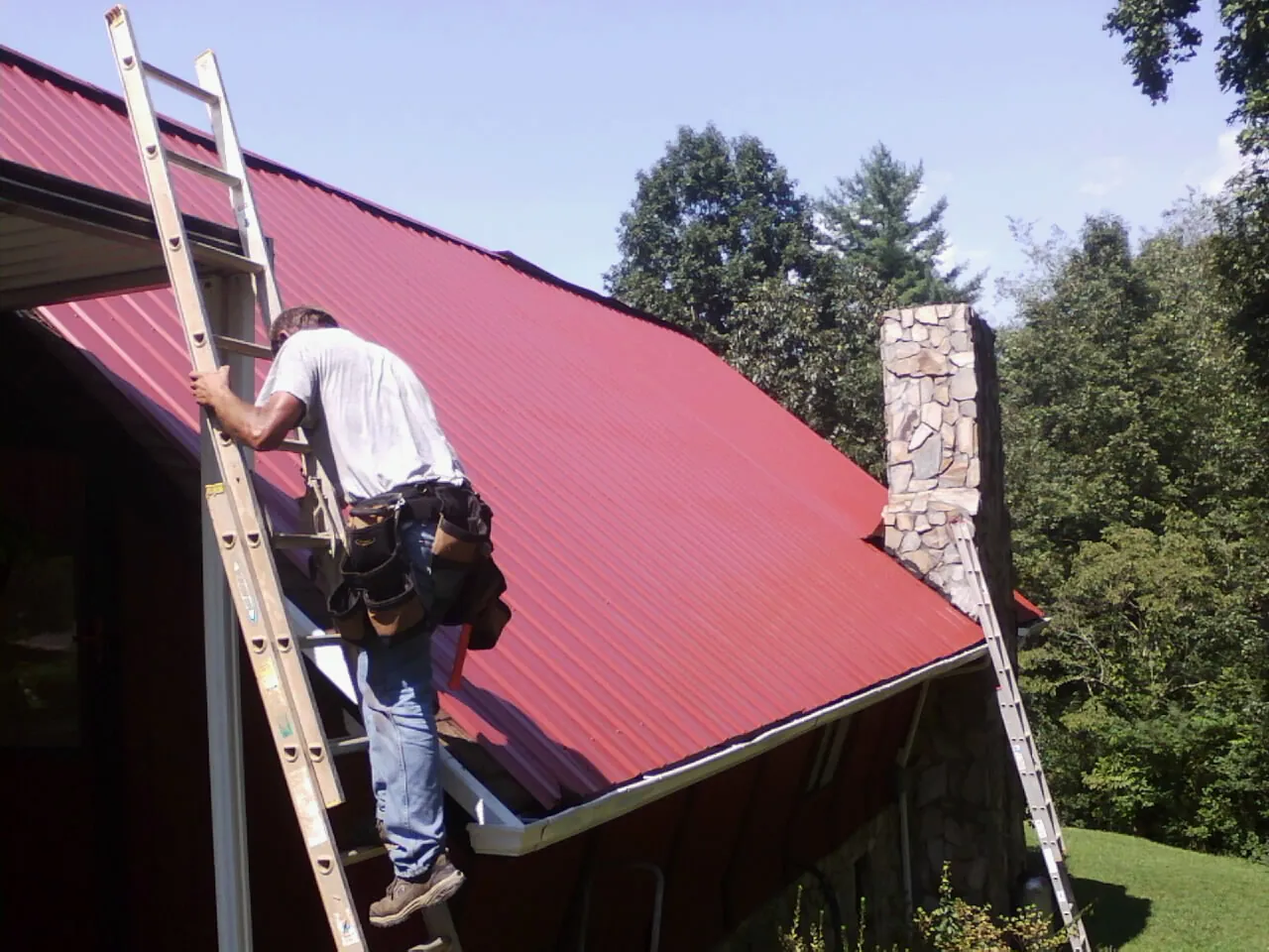 Roofer on a ladder beside a red metal roof and stone chimney — Soffit & Fascia Repair by Roofing Brothers in Mayodan